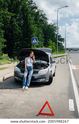 A young girl stands near a broken-down car in the middle of the highway and calls for help on the phone. Failure and breakdown of the car. Waiting for help.