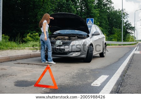 A young girl stands near a broken-down car in the middle of the highway and looks under the hood. Failure and breakdown of the car. Waiting for help.
