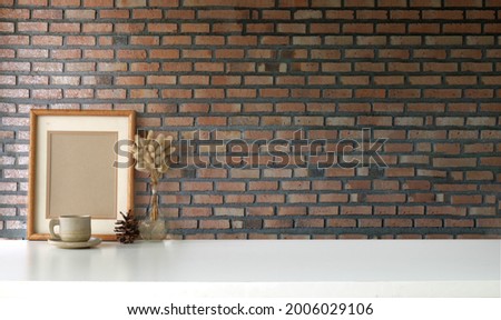 Empty picture frame and coffee cup on white table with brick wall. Copy space for graphics display montage.