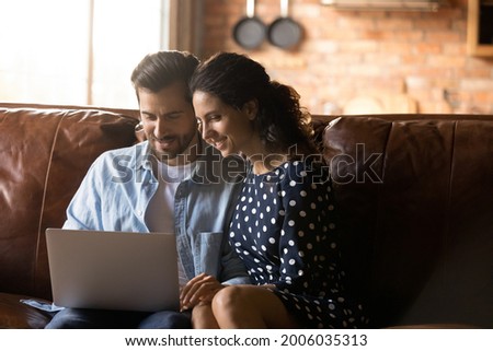 Happy couple sitting on couch at home together, using banking app on laptop, paying insurance, taxes, mortgage bills, planning family budget, making video call, shopping online, browsing internet