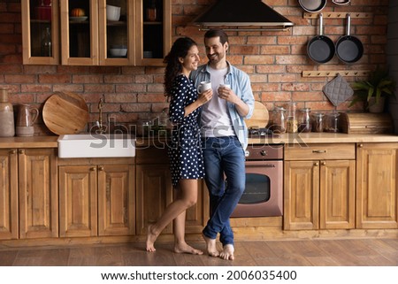 Happy sweet barefooted young couple drinking coffee in stylish kitchen together, holding mugs, hugging, talking, laughing, enjoying morning leisure time and hot beverage at home. Full length Royalty-Free Stock Photo #2006035400