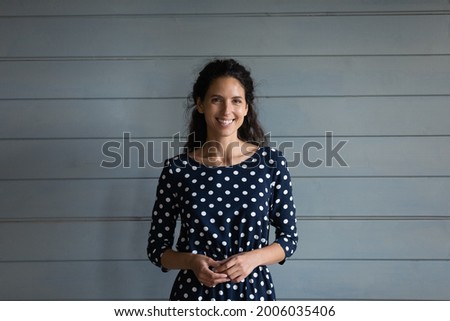 Portrait of happy beautiful Latin millennial 30s woman wearing spotted dress, standing against grey background with toothy smile, looking at camera and laughing. Head shot, profile picture