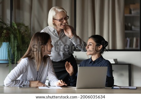 Diverse female office team discussing project and laughing. Business women of different ages and ethnicity talking at workplace with laptop, having fun, sharing good news, enjoying working together Royalty-Free Stock Photo #2006035634