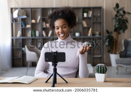 Smiling African American woman blogger influencer recording video on smartphone on tripod at workplace, businesswoman business coach mentor shooting webinar or online training, sitting at desk