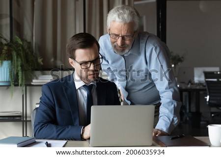 Older professional giving help, advice to young coworker, explaining issues, supervising work of new employee. Businessmen discussing project at laptop, looking at screen. Mentor training intern Royalty-Free Stock Photo #2006035934
