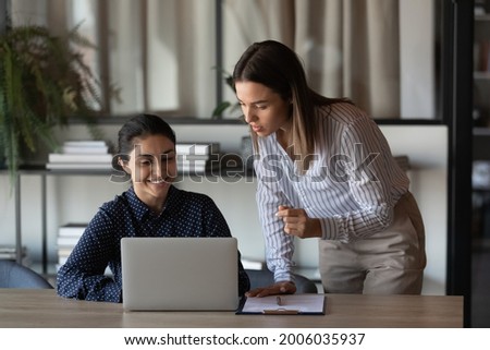 Happy Indian office employee showing job result to boss. Female coworkers discussing project at laptop, talking, laughing. Mentor training intern, supervising work of trainee, explaining task Royalty-Free Stock Photo #2006035937