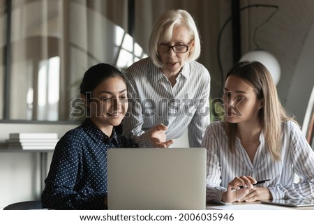 Corporate teacher showing and explaining work data to student girls, pointing at laptop screen, speaking. Mentor training interns. Elder coach giving advice to diverse office apprentices Royalty-Free Stock Photo #2006035946