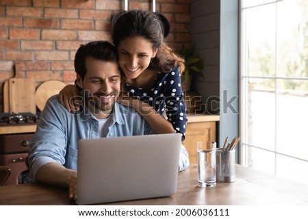 Happy millennial wife giving support to husband working at laptop from home, touching shoulders. Married couple surfing internet on computer together, paying bills online, reviewing mortgage terms Royalty-Free Stock Photo #2006036111