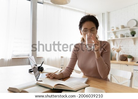Concentrated millennial female student in eyeglasses reading educational book, drinking glass of fresh pure water, staying refreshed while preparing for exams in modern kitchen, learning concept. Royalty-Free Stock Photo #2006039210