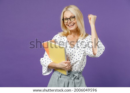 Happy gray-haired blonde teacher woman lady 40s 50s years old in white dotted blouse eyeglasses standing hold notepads doing winner gesture isolated on bright violet color background studio portrait