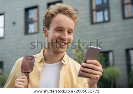 Handsome smiling man wearing yellow shirt using mobile phone, shopping online, reading good news standing on the street. Portrait of happy tourist holding smartphone searching way 