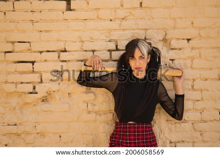Pretty young girl with heterochromia and punk style with a baseball bat back of her head resting on a yellow brick wall in the background.