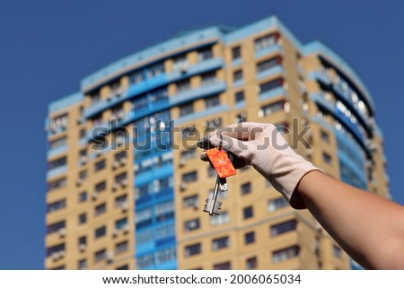 House purchase, woman in protective gloves holding keychain with digital and home keys on background of new buildings. Moving home or renting property during coronavirus pandemic