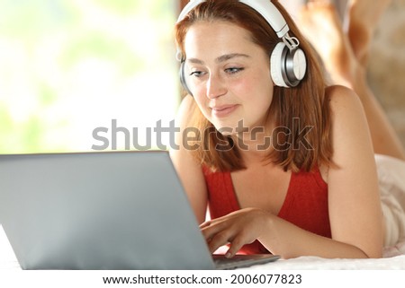Satisfied woman with wireless headphones checking laptop lying on the bed in the bedroom