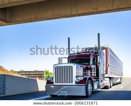 Red industrial long haul Big rig bonnet semi truck with chrome transporting frozen commercial cargo in refrigerator semi trailer running for delivery on wide multiline highway road under the bridge
 Royalty-Free Stock Photo #2006131871
