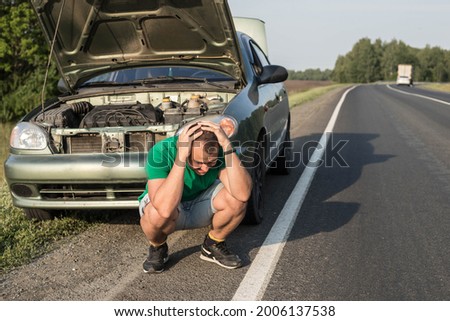 An unfortunate driver is in shock near a broken car in the middle of the highway. A broken car with an open hood on the side of the road with the driver clutching his head. Royalty-Free Stock Photo #2006137538