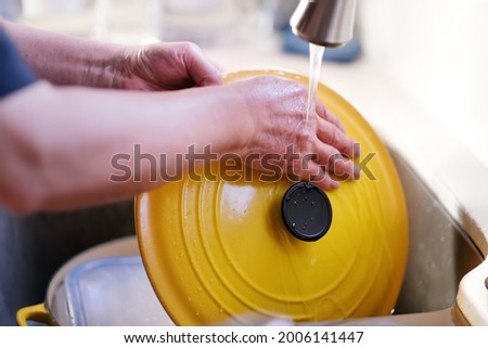 Man hands washing cleaning beautiful yellow cast iron cooking pot lid and dishes after dinner at modern kitchen sink in running water under chrome faucet. Environmental, water conservation and family.