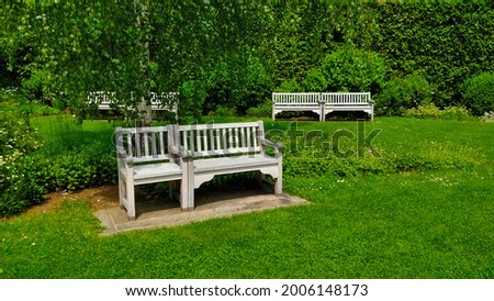 White bench in front of the image with two benches in the background. There is grass all over the picture and behind the seat is tall hedgerow.