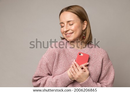 Happy pleased woman hug smartphone. Young blond female press mobile phone to chest get pleasant message from boyfriend lover with dreamy cute face closed eyes. Girl in love isolated over studio wall Royalty-Free Stock Photo #2006206865