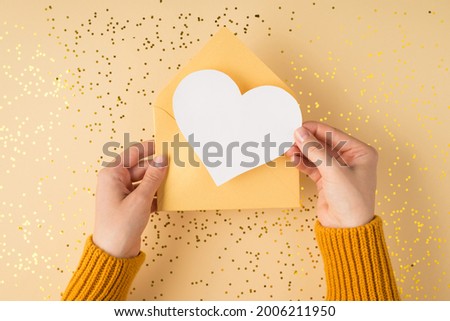 First person top view photo of female hands in yellow sweater holding open pastel yellow envelope with white paper heart over scattered golden sequins isolated light orange background with copyspace