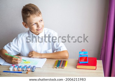 a blond boy in a white shirt draws in an album with a pencil against a background of paints, brushes, pencils and a red book, sitting at a desk at school