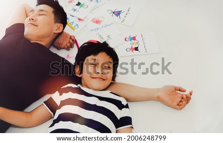 Top view cute father and son lying and pretending to sleep together on the white cement floor at home after finishing the greeting card with crayons : Two happy family between father and naughty son.