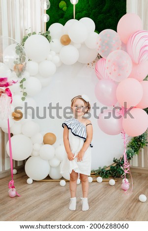 A little girl in a white dress stands on a photo zone with balloons for her birthday. Festive decor. happy and cheerful children's holiday.