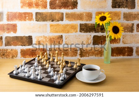 Interior image with sunflower flowers, coffee cups and chess board on the table in the room with brick walls.