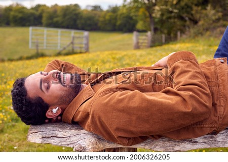 Man Lying On Bench In Countryside Relaxing And Listening To Music Or Podcast On Wireless Earphones