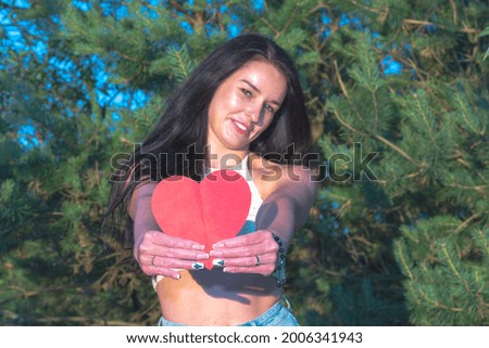 Pretty brunette showing red heart postcard smiling dreamy nature summer sunny day.Woman in nature holding a red paper heart in front.Green background.Selective focus.