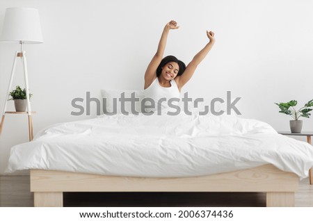 Good start of new day. Well-slept african american lady stretching arms after waking up, feeling happy and full of vitality. Young woman sitting in comfortable bed in coy bedroom interior Royalty-Free Stock Photo #2006374436