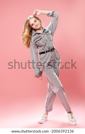 Full length portrait of a smiling girl teenager posing in fashionable denim overalls on a pink background. Studio fashion shot. Youth style. 