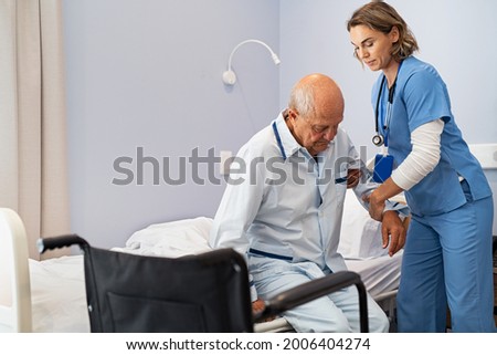 Lovely nurse assisting senior man to get up from bed at hospital. Caring nurse supporting elderly patient while getting up from bed and move towards wheelchair at nursing home.  Royalty-Free Stock Photo #2006404274