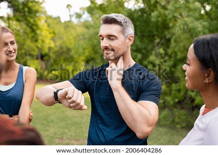 Mature tired man checking pulse after workout. Handsome middle aged man standing with group of friends measuring heart rate pulse on his neck and looking sport watch. Guy times the pulsations at park. Royalty-Free Stock Photo #2006404286