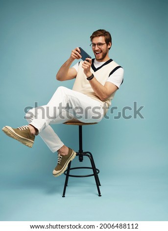 Portrait of an excited young man sitting on a stool and playing games on mobile phone isolated over blue background. Royalty-Free Stock Photo #2006488112