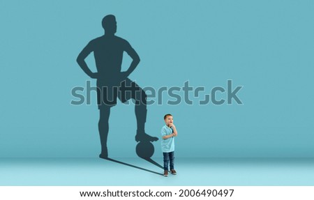 Childhood and dream about big and famous future. Conceptual image with boy and shadow of sportive male football player, champion on blue background. Childhood, dreams, imagination, education concept. Royalty-Free Stock Photo #2006490497
