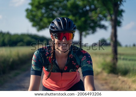 Close up Frontal photo of a young woman cyclist, smiling, riding her road bike, in the middle of nature, illuminated by sunlight. She is wearing a helmet, sunglasses, and a pink cycling outfit. Royalty-Free Stock Photo #2006493206