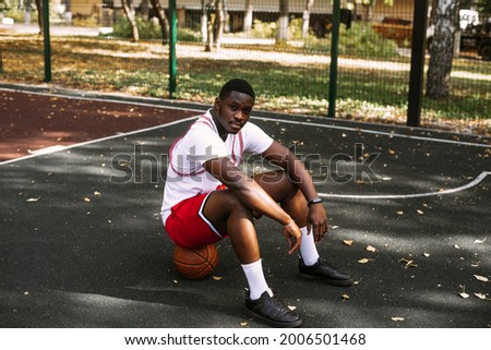 Portrait of an African American young man holding a basketball and sitting on a basketball court. Take a break during your workout. Fashionable sports portrait