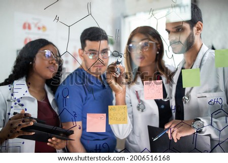 Medicine, technology, research concept. Team of four multiracial focused medical scientists working and writing on the glass board in modern office. Focus on hand of afro woman writing formula