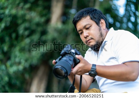 Asian beard Thai professional camera man check image shot via liveview behind Medium format Mirrorless camera with green tree background.