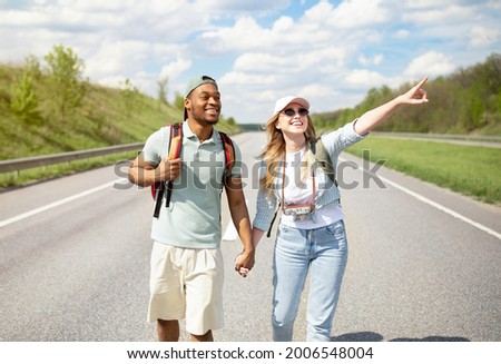 Cool young multiethnic couple holding hands while walking along highway with backpacks, hitchhiking together. African American guy and his girlfriend traveling by autostop