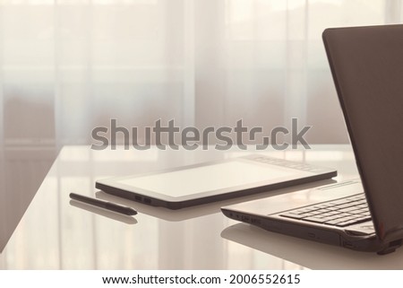 A graphic tablet and a laptop are lying on a glass table.