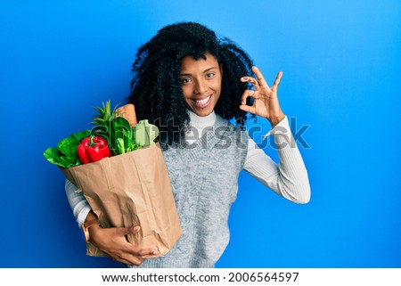 African american woman with afro hair holding paper bag with groceries doing ok sign with fingers, smiling friendly gesturing excellent symbol 