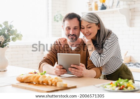 Mature middle-aged family couple wife and husband parents using digital tablet while having breakfast in the kitchen at home, reading news, searching web, reading e-book, e-learning remotely.