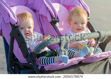 Two twin sisters are sitting next each other in a baby stroller with toys in their hands.