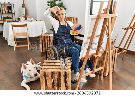 Caucasian young blonde woman sitting on wheelchair at art studio with angry face, negative sign showing dislike with thumbs down, rejection concept 