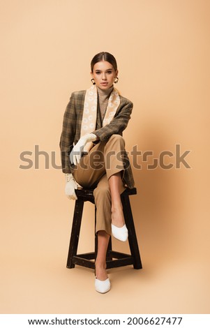 elegant woman in trousers, blazer, white shoes and gloves sitting on beige background Royalty-Free Stock Photo #2006627477