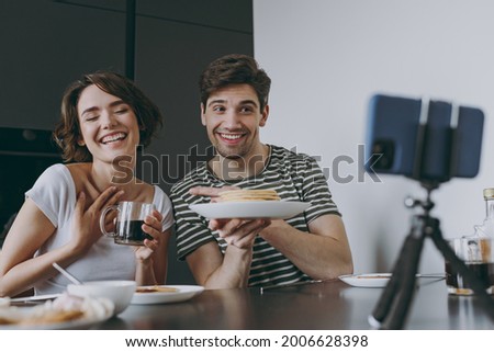 Young happy fun couple two woman man in casual clothes sit by table eat showing pancake talk video call mobile cell phone drink coffee cook food in light kitchen at home together Healthy diet concept