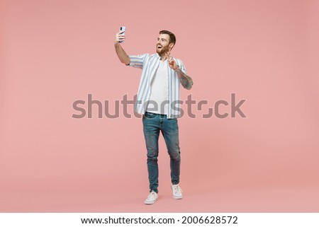 Full length young caucasian unshaven man in blue striped shirt doing selfie shot on mobile phone post photo on social network waving hand isolated on pastel pink background. People lifestyle concept