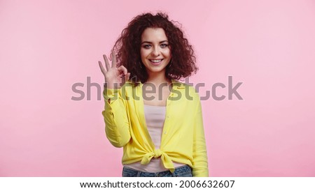 pleased young and curly woman showing ok sign isolated on pink